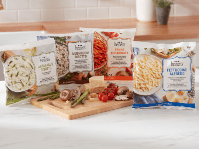 Heinen's Frozen Entrees on a counter next to a cutting board with some fresh cut veggies and cheese