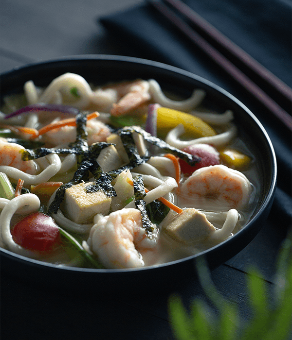 UDON NOODLES WITH SHRIMP, TOFU & VEGE TABLES served in a black bowl