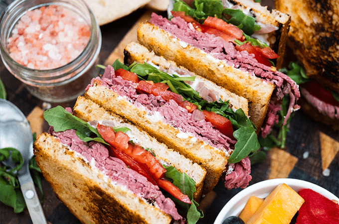 Yoko's Roast Beef & Arugula Sandwich stacked high and served with some fruit on a wooden cutting board