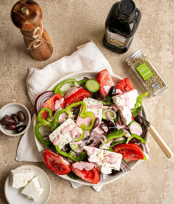 Traditional Greek Salad in a white bowl with a fork in it surrounded by its ingredients on a table