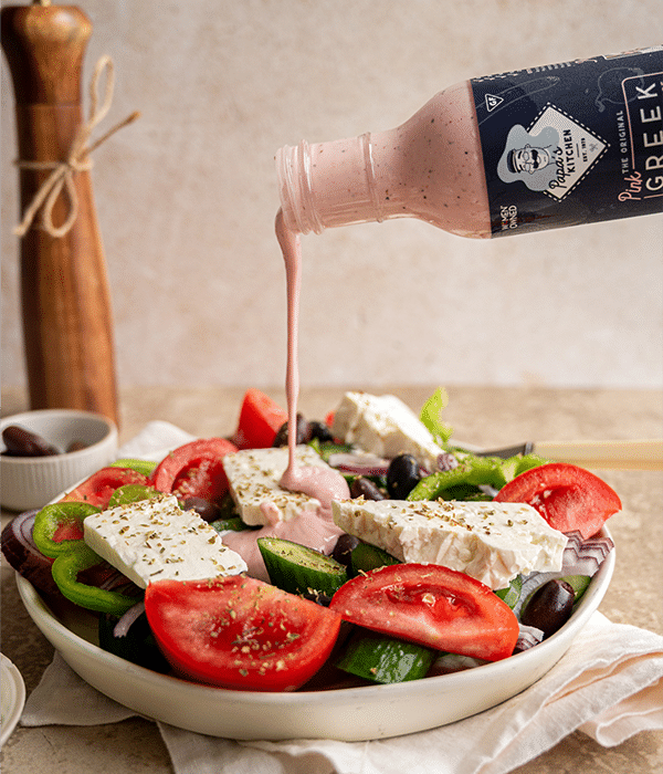 Traditional Greek Salad in a white bowl with dressing being poured into it