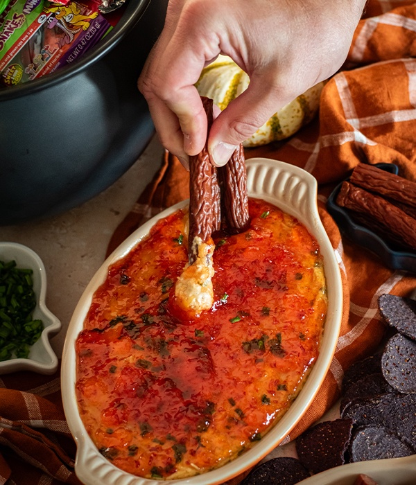A hand holding meat sticks, dipping them into a baked dish of pepper jelly cream cheese dip.
