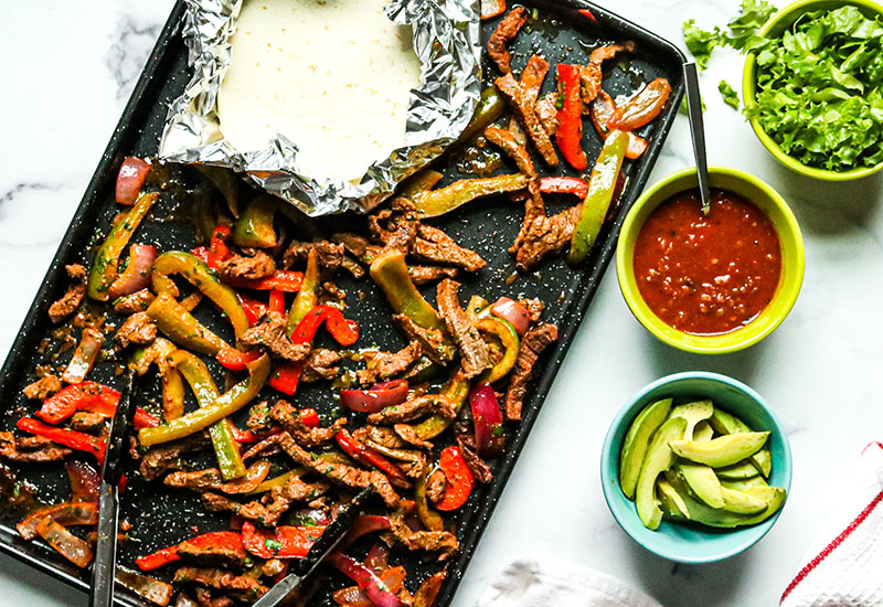 Cooked Fajita Meat and Vegetables on a Sheet Pan with Warm Tortillas and Sides of Avocado, Salsa and Greens