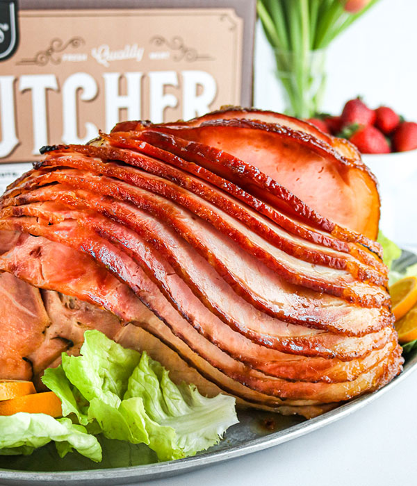 Heinen's Spiral Sliced Ham on a Platter with Fresh Greens. A Heinen's Butcher Shop Box and a Vase of Tulips are in the Background