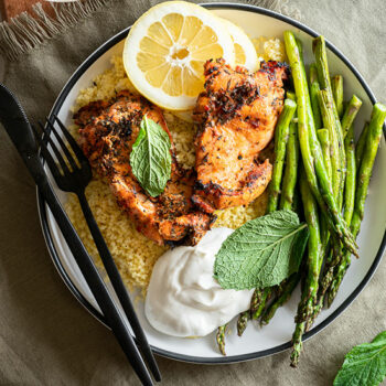 Heinen's Grilled Moroccan Chicken Thighs on a Plate with Rice. Asparagus and Lemon. A Package of Heinen's Marinated Chicken Thighs Sits Beside the Plate