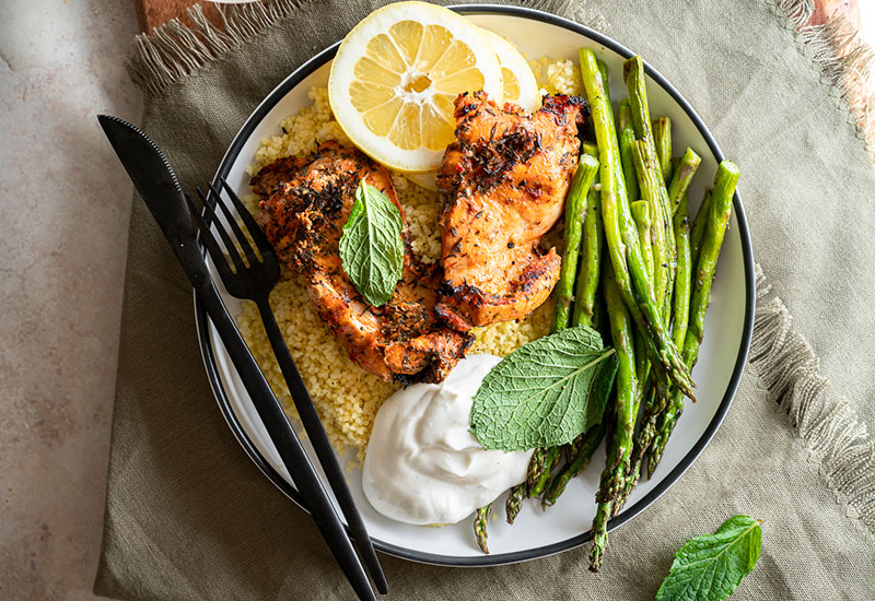 Heinen's Grilled Moroccan Chicken Thighs on a Plate with Rice. Asparagus and Lemon. A Package of Heinen's Marinated Chicken Thighs Sits Beside the Plate