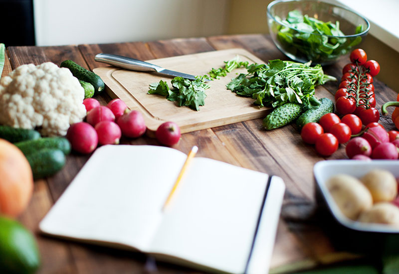 Fresh Fruits and Vegetables on a Countertop with a Cutting Board and a Notebook