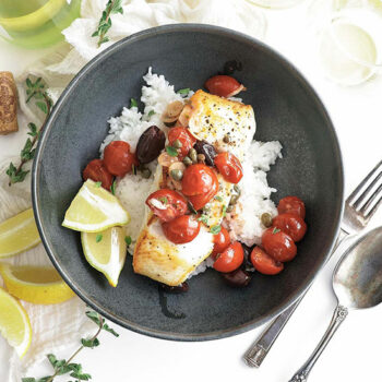 Mediterranean Halibut in a Bowl with Rice, Cherry Tomatoes, Olives and Lemon. Lemon Slices and a Glass of Wine on the Side