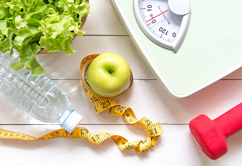 Weight Scale on a Light-Colored Surface with Measuring Tape, Leafy Greens, An Apple, A Water Bottle and a Hand Weight