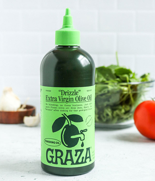 Graza Drizzling Oil in its bottle on a on a Slate Gray Surface with a subway tile background, a bowl of greens, a tomato and bulbs of garlic.