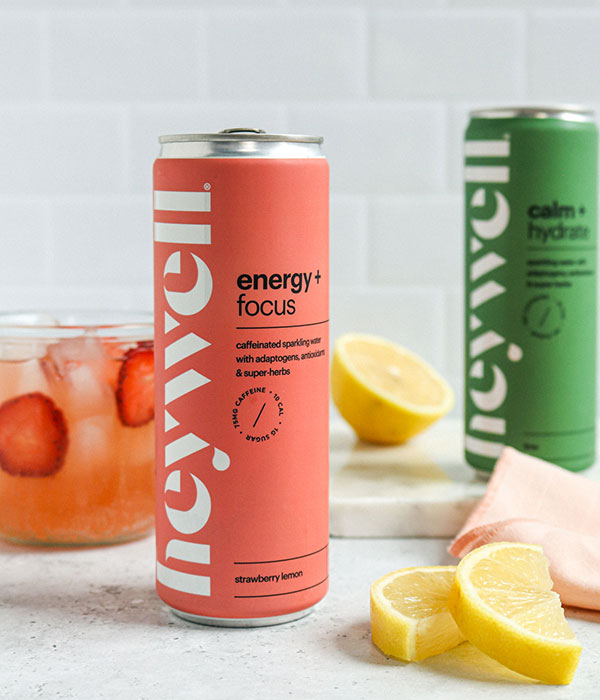 Heywell Sparkling Water cans on a Slate Gray Surface with a subway tile background, fresh slices lemon and a strawberry drink in a glass.