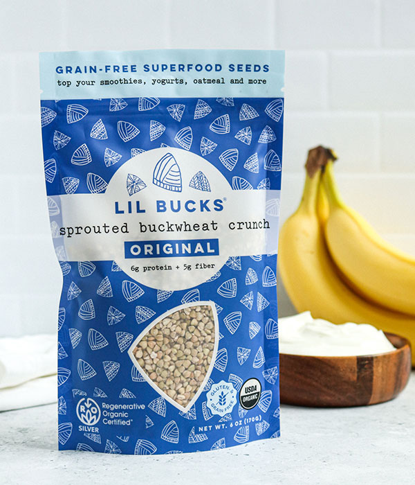 Lil Bucks Buckwheat Granola in its packaging on a on a Slate Gray Surface with a subway tile background, bananas and a bowl of coconut flakes.