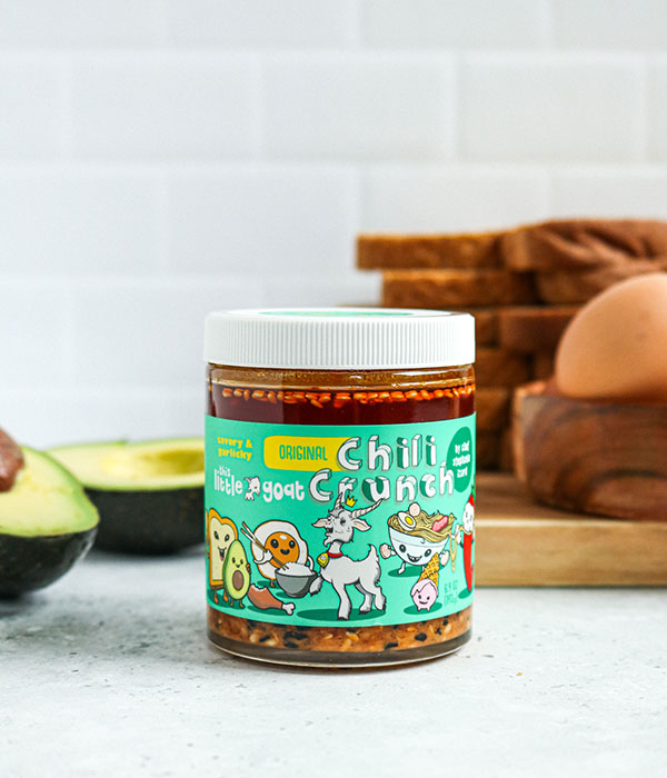 This Little Goat Chili Crunch Oil in its packaging on a on a Slate Gray Surface with a subway tile background, bread slices and fresh avocados.