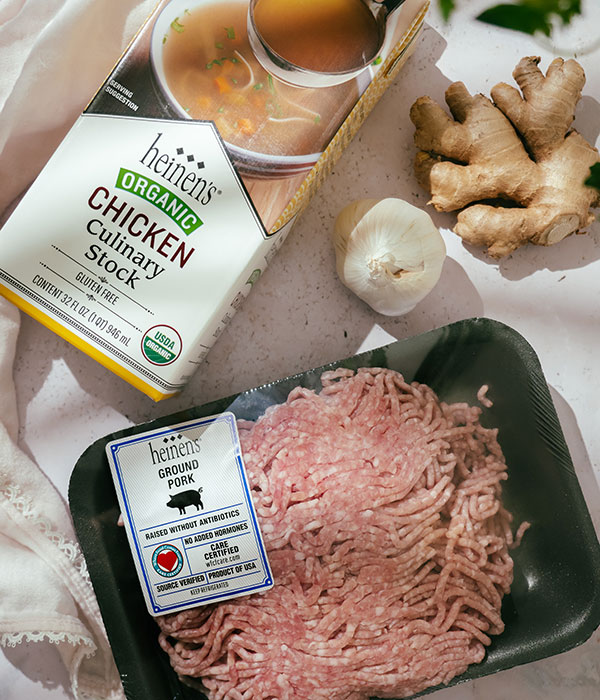 Heinen's Raw CARE Certified Ground Pork in Packaging. Heinen's Chicken Stock Packaging, Fresh Garlic Bulb and Fresh Ginger Laying Flat on a Surface.