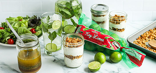 Fresh Key Limes in a Bag on a Light Surface surrounded by a Citrus Marinade, Fresh Green Salad, Yogurt and Granola Parfaits and Fresh Granola on a Sheet Tray. 