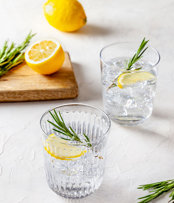 Twi Glasses of Ice Water with Fresh Slices of Lemon and Fresh Sprigs of Rosemary. Fresh Rosemary and Fresh Lemons are in the background