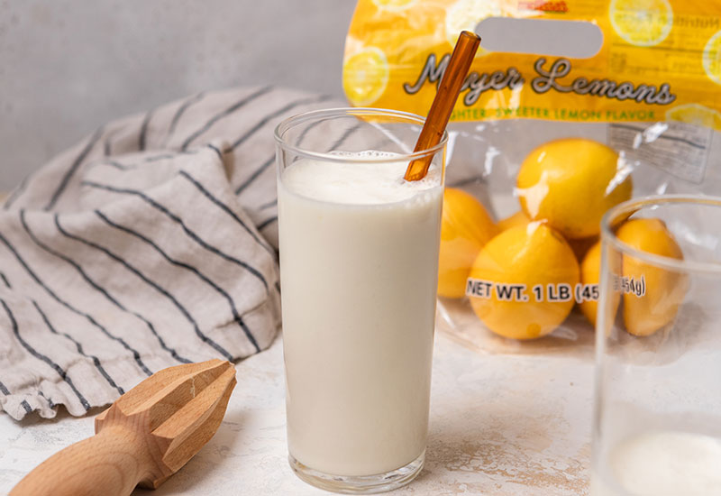 Copycat Frosted Lemonade in a Glass with a Straw, Dish Towel and a bag of Meyer Lemons in the Background.
