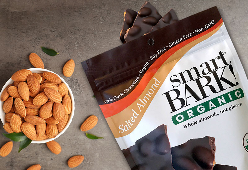 An open bag of salted almond smartBARK! on a neutral surface with a bowl of almonds