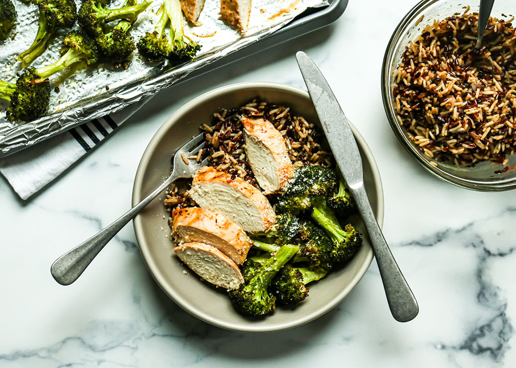 Heinen's Cooked Marinated Sundried Tomato Chicken in a Bowl with Rice and Broccoli/ A Bowl of Rice and a sheet pan of broccoli is surrounding the bowl