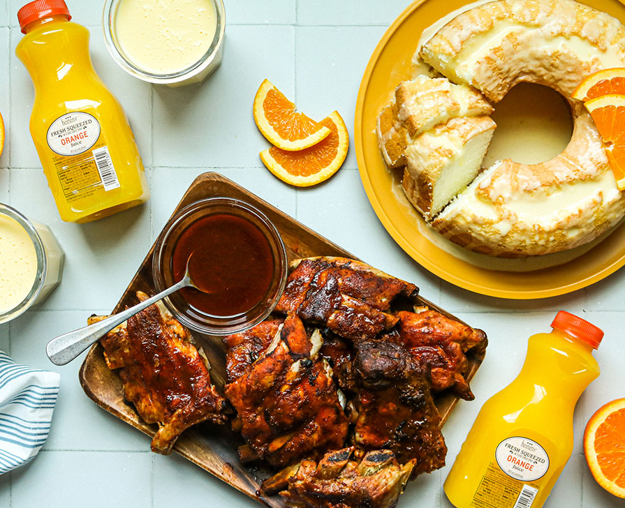Citrus Glazed Ribs and a Citrus Glazed Angel Food Cake on a Tile Surface with Containers of Heinen's Fresh Squeezed Orange Juice and Fresh Oranges Slices.