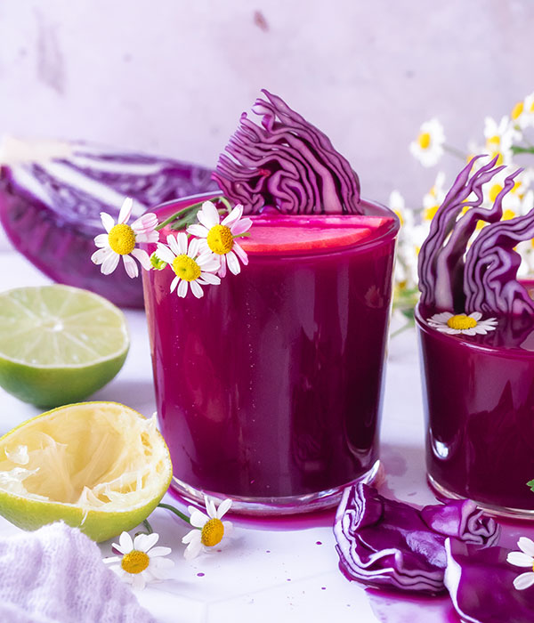 A Glass of Purple Power Juice. Fresh Cabbage and Halved Limes Beside the Glass.