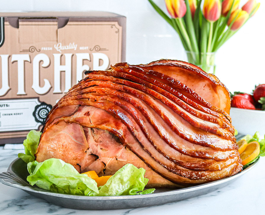Heinen's Full Spiral Sliced Ham on a Serving Plate with Fresh Lettuce and Orange Slices. A Vase of Tulips and Heinen's Butcher Shop Box Sits in the Background. 