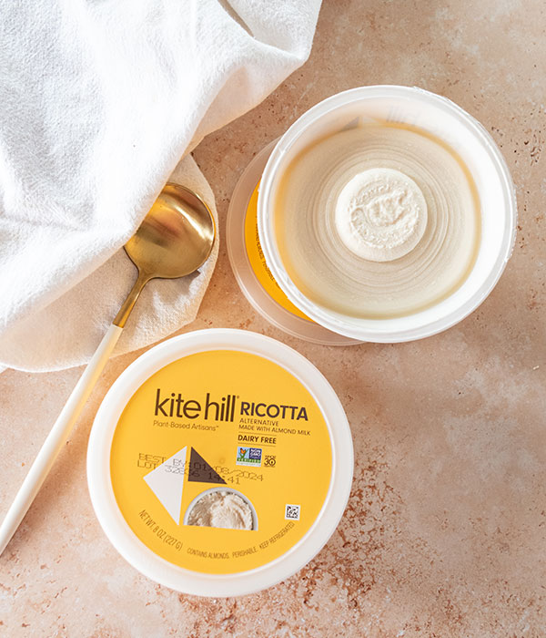 Kite Hill Ricotta Packaging on a Neutral Surface with a Spoon and White Dish Towel