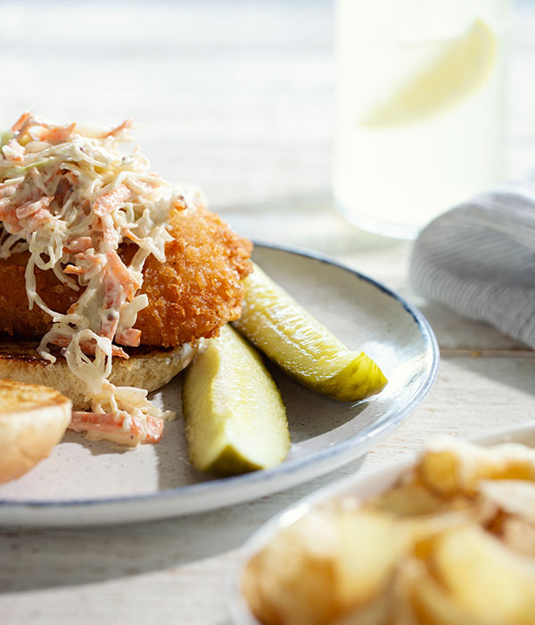 Vertical Image of a Lemon Slaw Fish Sandwich on a Plate with Pickle Spears, a Bowl of Kettle Chips and a Glass of Water