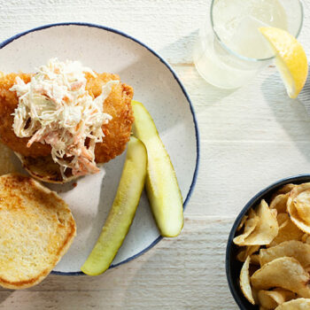 Horizontal Image of a Lemon Slaw Fish Sandwich on a Plate with Pickle Spears, a Bowl of Kettle Chips and a Glass of Water