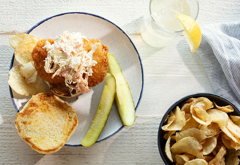 Horizontal Image of a Lemon Slaw Fish Sandwich on a Plate with Pickle Spears, a Bowl of Kettle Chips and a Glass of Water