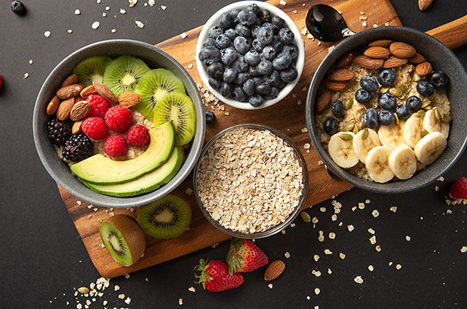 Top Down Image of Two Oatmeal Bowls with Fresh Fruit and Nut Toppings. Bowls of oats and blueberries beside.