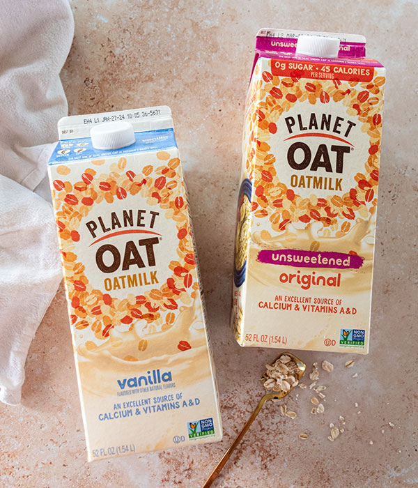 Planet Oat Oat Milk Cartons on a Neutral Surface with a White Dish Towel, Gold Spoon and Loose Oats