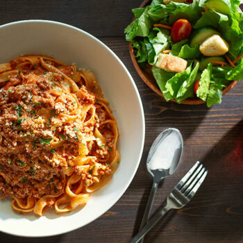 Horizontal Top Down Image of Simple Fettuccine Bolognese in a Bowl with a Side Salad, a Glass of Red Wine and a Spoon and Fork