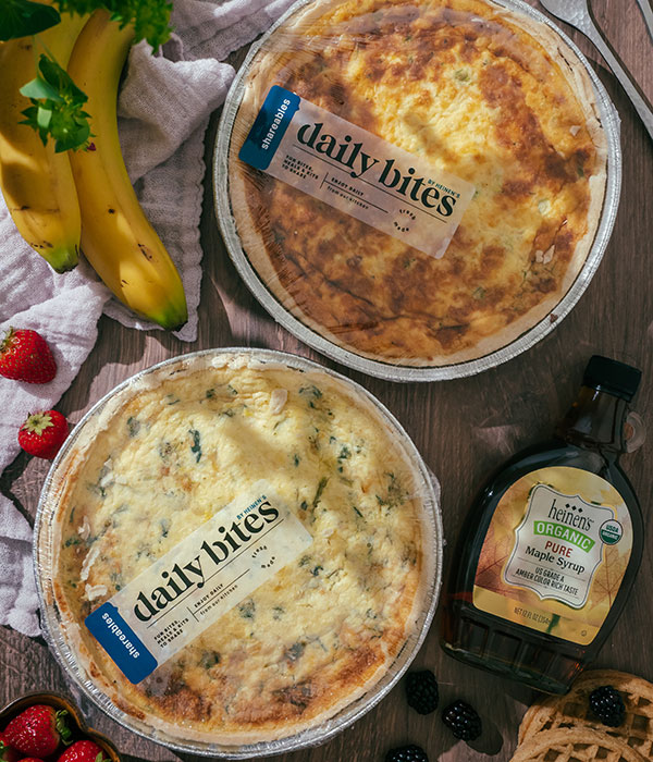 Two Heinen's Daily Bites Quiches in their packaging with Heinen's Maple Syrup and Fresh Fruits and Vegetables