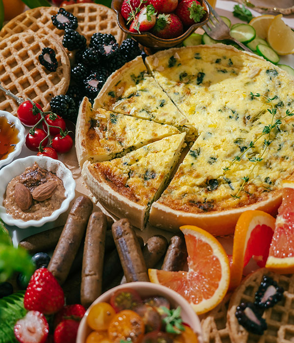 Assortment of Brunch Food Including Sliced Quiche, Breakfast Sausage, Toaster Waffles and Fresh Fruit and Vegetables