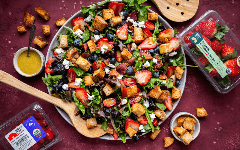 Bacon Berry Salad in large serving bowl with croutons, tomatoes, and strawberries laid out around it.