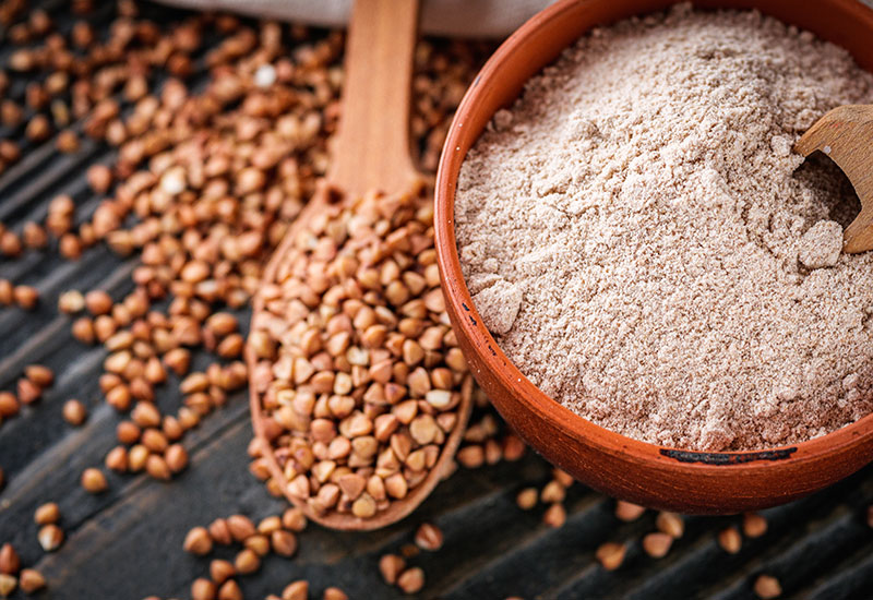 A Wooden Spoon with Buckwheat Seeds Beside a Bowl of Buckwheat Flour