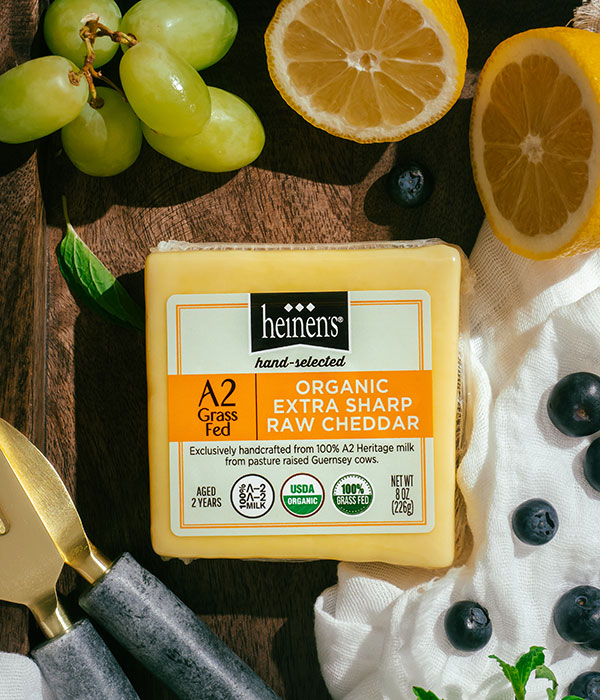 Heinen's Hand-Selected A2 Organic Extra Raw Sharp Cheddar Cheese on a White Serving Cloth and Serving Board with Fresh Lemons, Fresh Blueberries, Fresh Herbs, Green Grapes and Cheese Servingware