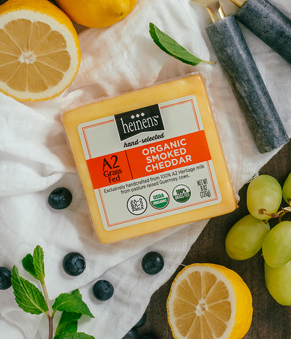 Heinen's Hand-Selected A2 Organic Smoked Cheddar Cheese on a White Serving Cloth with Fresh Lemons, Fresh Blueberries, Fresh Herbs, Green Grapes and Cheese Servingware
