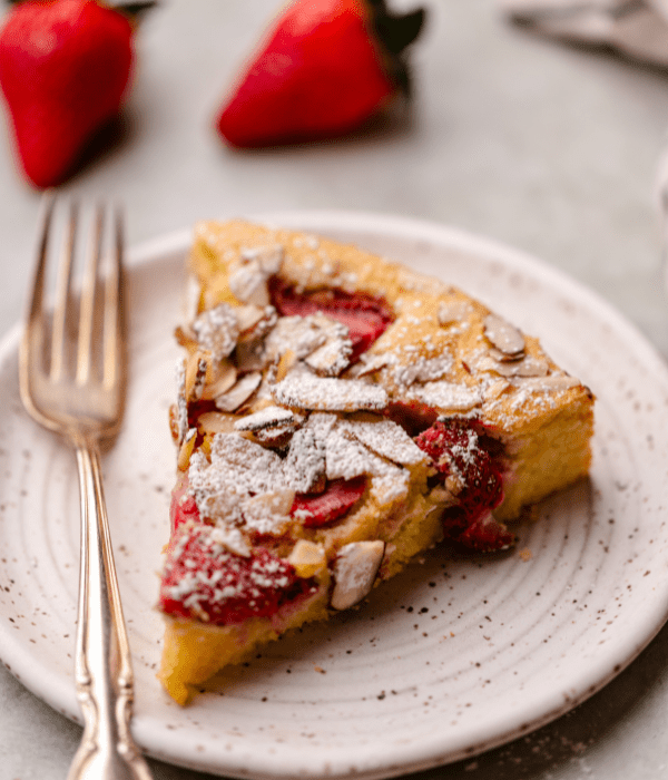Slice of Strawberry Almond cake on white plate with fork on a table with strawberries in the background 